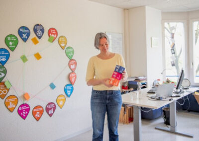 Frau steht in einem Büro. An der Wand hängen die 17 SDG-Icons, die Frau stapelt 3 SDG-Würfel zwischen ihren Händen.